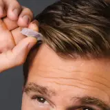 Mann mit beginnenden Geheimratsecken, der eine kleine Kapsel ins Haar drückt.