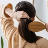 Frau bürstet ihr langes, dunkles Haar mit einer Holzbürste. Sie trägt einen Bademantel. Ein Spiegel zeigt sie lächelnd. So kann man **Haare schneller wachsen lassen**.