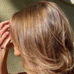 Glänzendes, welliges Haar in warmer bronde haarfarbe, das im Sonnenlicht schimmert. Eine Hand streicht sanft durch die Strähnen.