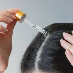 Eine Hand trägt mit einer Pipette eine Flüssigkeit auf die Kopfhaut auf. Ein Tropfen fällt auf den Scheitel. Apfelessig gegen Schuppen kann hier helfen.