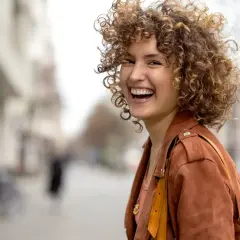 Lachende Frau mit voluminösem, lockigem Haar in brauner Lederjacke auf einer belebten Straße.