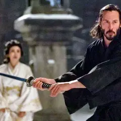 Ein Mann mit langer, dunkler Samurai-Frisur und Bart hält ein Schwert. Im Hintergrund eine Frau in weißem Kimono.