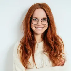 Eine lächelnde Frau mit langen, roten Haaren und Brille. Ihr Outfit, ein cremefarbener Strickpullover, steht ihr gut.