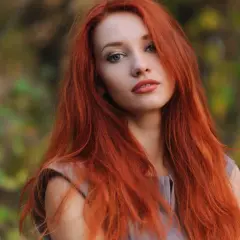 Eine Frau mit langen, dunkelroten Haaren blickt nachdenklich in die Kamera. Ihr Haar fällt ihr über die Schultern, während sie im Freien steht.