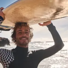 Lächelnder Surfer mit Wellen im Haar trägt sein Brett über dem Kopf. Sein Neoprenanzug hat ein cooles Schachbrettmuster.