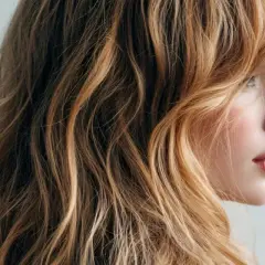 Profil einer Frau mit welligem, honigblondem Haar und zarten, wispy bangs, die ihr Gesicht umspielen.