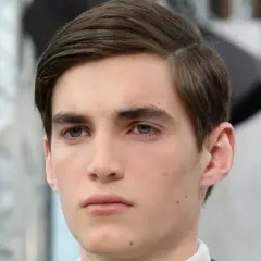 Ein junger Mann mit braunem Haar, das zu einem perfekten Seitenscheitel gestylt ist. Sein Blick ist nachdenklich.