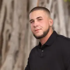 Junger Mann mit Bart und Buzz Cut 10mm, trägt schwarzes Poloshirt, steht vor unscharfem Baumstamm.