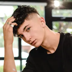 Junger Mann mit kurzgeschorenen Seiten und einem kurzen Mullet, der nachdenklich seine Hand an den Kopf legt.