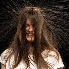 Lachende Frau mit Haaren, die elektrisch abstehen. Eine Bürste in der Hand verstärkt den Eindruck von statischer Aufladung.