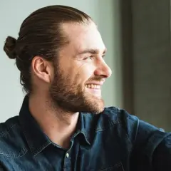 Lächelnder Mann mit Bart und einem lässigen **man bun** im Jeanshemd.