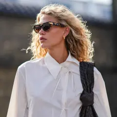 Eine skandinavische Blonde mit lockigem Haar, Sonnenbrille und weißem Hemd, die eine dunkle Tasche trägt.