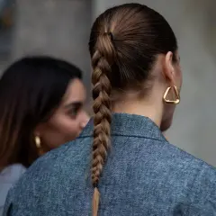Eine Frau mit geflochtenen Haaren trägt einen grauen Blazer und auffällige goldene Ohrringe.