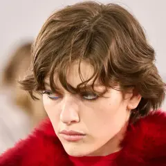 Ein Model mit modernen Kurzhaarfrisuren und auffälligem blauen Eyeliner, gekleidet in einen roten Pelzmantel.