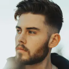 Junger Mann mit Bart und einem modernen taper fade cut, der nachdenklich in die Ferne blickt.