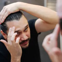 Mann prüft seine Haare im Spiegel. Er überlegt, ob Aminexil oder Minoxidil besser gegen Haarausfall hilft.
