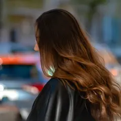 Braune Haare mit Beach Waves, die im Wind wehen. Die Frau trägt eine schwarze Lederjacke. Im Hintergrund verschwommene Autos.