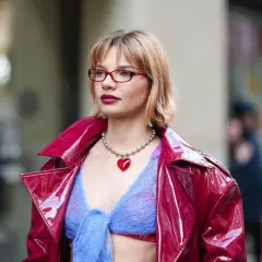 Frau mit roter Brille und einem roten Herz-Anhänger trägt eine glänzende rote Jacke über einem blauen Strickoberteil. Ihr **bob hinten kurz gestuft** umrahmt ihr Gesicht.