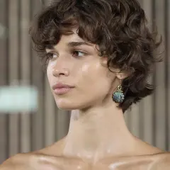 Ein Porträt einer Frau mit kurzen Haaren, die in natürlichen Locken fallen. Ihr Blick ist nach links gerichtet, und sie trägt einen auffälligen Ohrring.