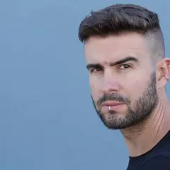 Junger Mann mit kurz geschorenen Seiten und Haare nach hinten, Bart und Lippenpiercing.