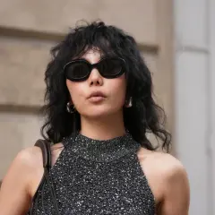 Frau mit schwarzem, gewelltem Haar im **Wolf Cut kurz**. Sie trägt eine glitzernde Top und eine Sonnenbrille.
