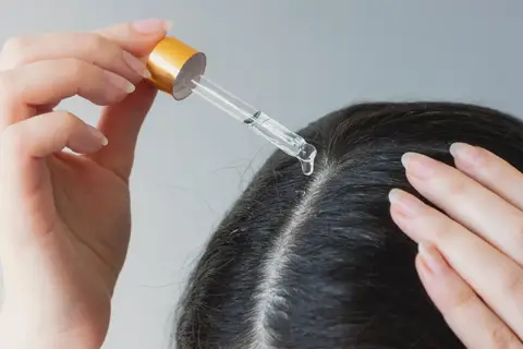 Eine Hand trägt mit einer Pipette eine Flüssigkeit auf die Kopfhaut auf. Ein Tropfen fällt auf den Scheitel. Apfelessig gegen Schuppen kann hier helfen.