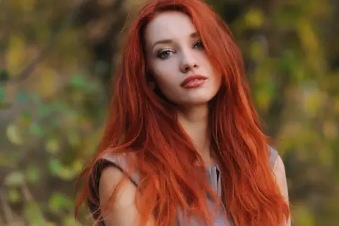 Eine Frau mit langen, dunkelroten Haaren blickt nachdenklich in die Kamera. Ihr Haar fällt ihr über die Schultern, während sie im Freien steht.