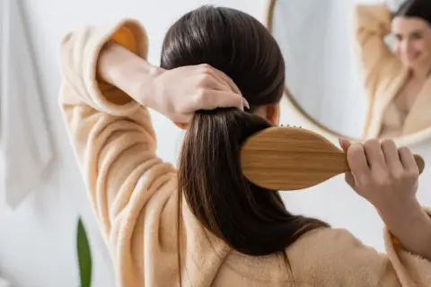 Frau bürstet ihr langes, dunkles Haar mit einer Holzbürste. Sie trägt einen Bademantel. Ein Spiegel zeigt sie lächelnd. So kann man **Haare schneller wachsen lassen**.