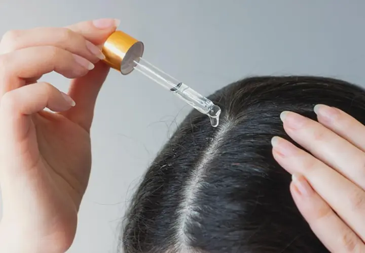 Eine Hand trägt mit einer Pipette eine Flüssigkeit auf die Kopfhaut auf. Ein Tropfen fällt auf den Scheitel. Apfelessig gegen Schuppen kann hier helfen.