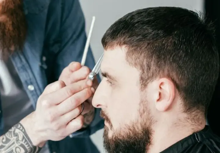 Ein Mann bekommt einen Caesar Cut. Der Friseur schneidet ihm mit einer Schere die Haare.