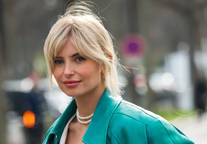 Blonde Frau mit soft curtain bangs, trägt eine türkise Lederjacke und Perlenkette.