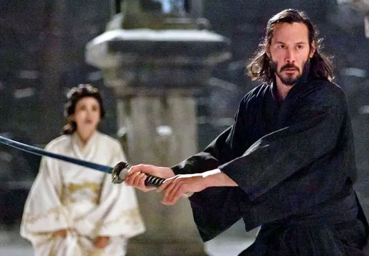 Ein Mann mit langer, dunkler Samurai-Frisur und Bart hält ein Schwert. Im Hintergrund eine Frau in weißem Kimono.