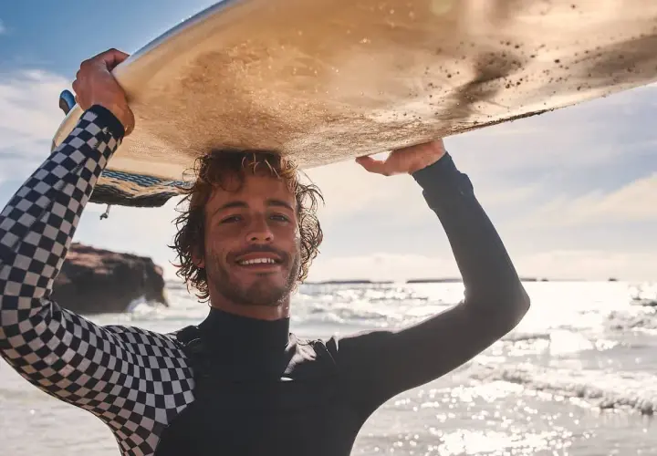 Lächelnder Surfer mit Wellen im Haar trägt sein Brett über dem Kopf. Sein Neoprenanzug hat ein cooles Schachbrettmuster.