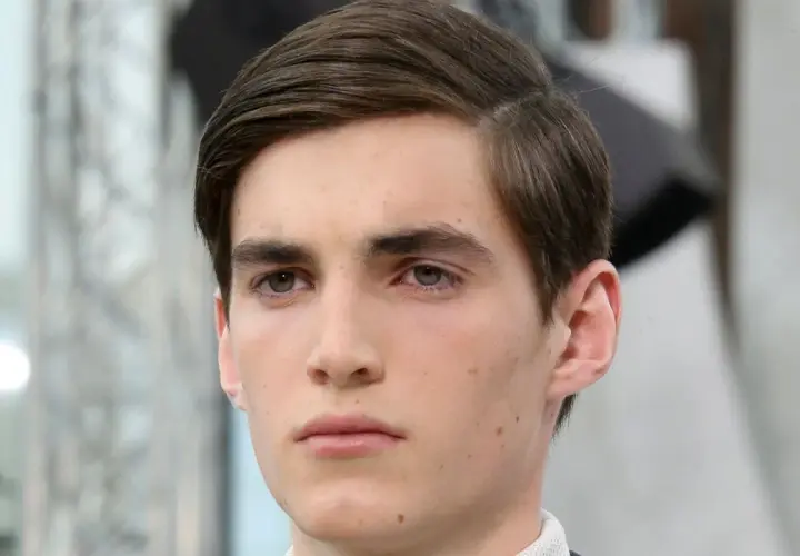 Ein junger Mann mit braunem Haar, das zu einem perfekten Seitenscheitel gestylt ist. Sein Blick ist nachdenklich.