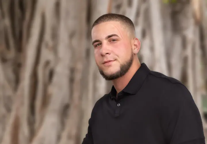 Junger Mann mit Bart und Buzz Cut 10mm, trägt schwarzes Poloshirt, steht vor unscharfem Baumstamm.