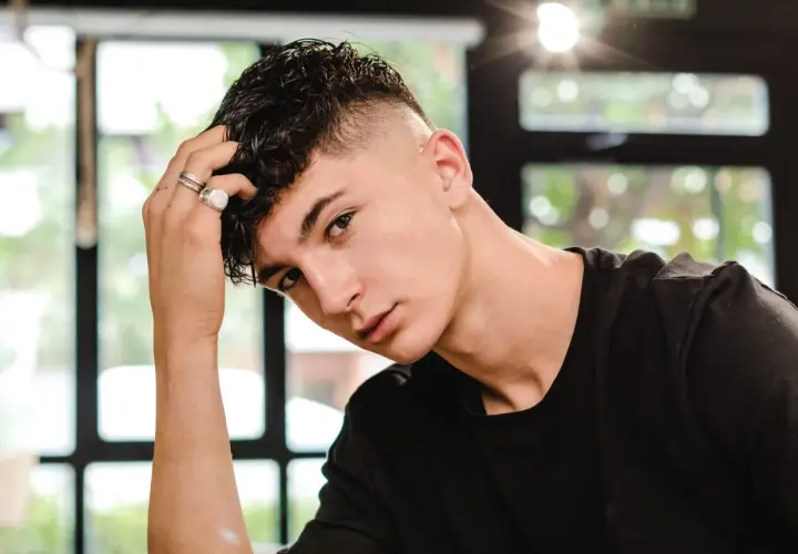 Junger Mann mit kurzgeschorenen Seiten und einem kurzen Mullet, der nachdenklich seine Hand an den Kopf legt.