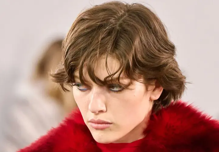 Ein Model mit modernen Kurzhaarfrisuren und auffälligem blauen Eyeliner, gekleidet in einen roten Pelzmantel.