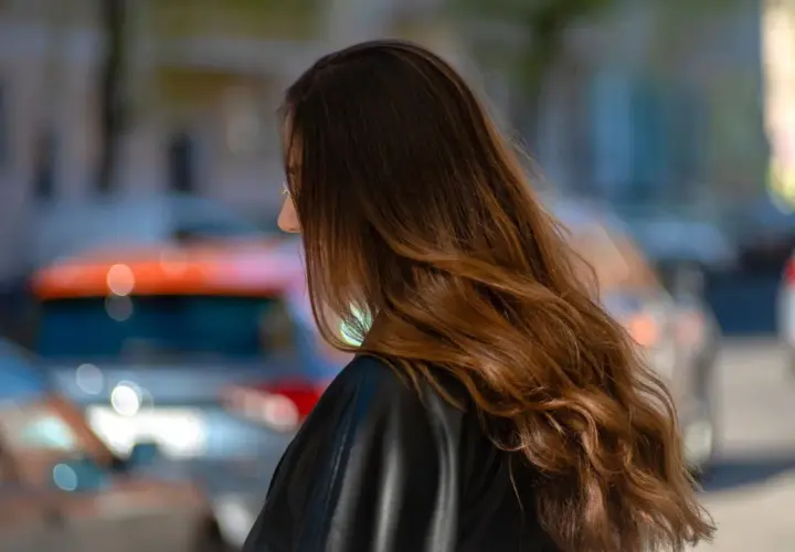 Braune Haare mit Beach Waves, die im Wind wehen. Die Frau trägt eine schwarze Lederjacke. Im Hintergrund verschwommene Autos.