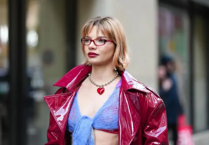 Frau mit roter Brille und einem roten Herz-Anhänger trägt eine glänzende rote Jacke über einem blauen Strickoberteil. Ihr **bob hinten kurz gestuft** umrahmt ihr Gesicht.