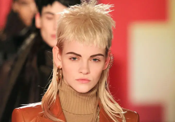 Ein Model präsentiert einen modernen Mullet mit blonden Locken, auffälligen Ohrringen und einem braunen Rollkragenpullover.