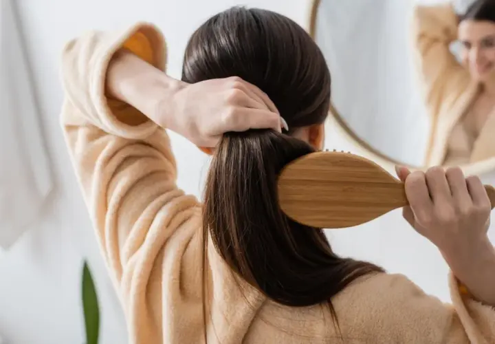 Frau bürstet ihr langes, dunkles Haar mit einer Holzbürste. Sie trägt einen Bademantel. Ein Spiegel zeigt sie lächelnd. So kann man **Haare schneller wachsen lassen**.
