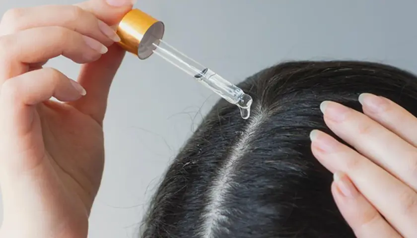 Eine Hand trägt mit einer Pipette eine Flüssigkeit auf die Kopfhaut auf. Ein Tropfen fällt auf den Scheitel. Apfelessig gegen Schuppen kann hier helfen.