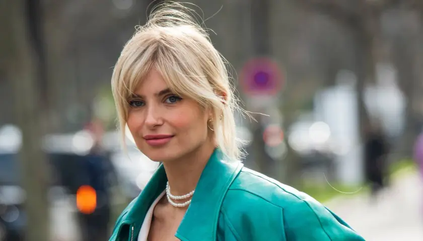 Blonde Frau mit soft curtain bangs, trägt eine türkise Lederjacke und Perlenkette.