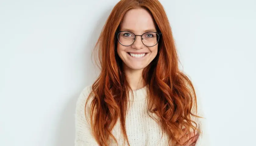 Eine lächelnde Frau mit langen, roten Haaren und Brille. Ihr Outfit, ein cremefarbener Strickpullover, steht ihr gut.