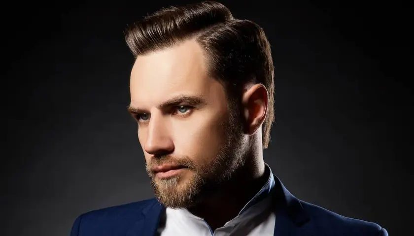 Ein Mann mit Bart und einem eleganten Seitenscheitel, gekleidet in einem blauen Anzug.