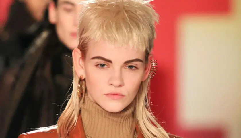 Model mit modernem Mullet-Haarschnitt, braunem Rollkragenpullover und Lederjacke. Goldene Ohrringe und auffälliger Ohrschmuck.