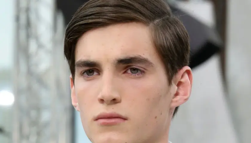 Ein junger Mann mit braunem Haar, das zu einem perfekten Seitenscheitel gestylt ist. Sein Blick ist nachdenklich.