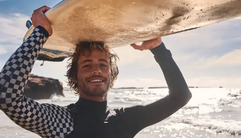 Lächelnder Surfer mit lockiger surfer frisur trägt Neoprenanzug und hält Surfbrett über dem Kopf.