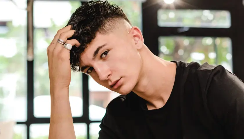 Junger Mann mit kurzgeschorenen Seiten und einem kurzen Mullet, der nachdenklich seine Hand an den Kopf legt.