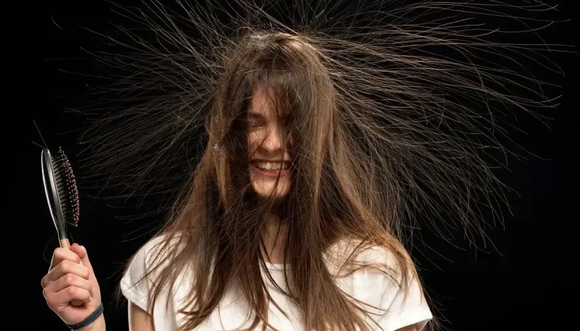 Lachende Frau mit Haaren, die elektrisch abstehen. Eine Bürste in der Hand verstärkt den Eindruck von statischer Aufladung.